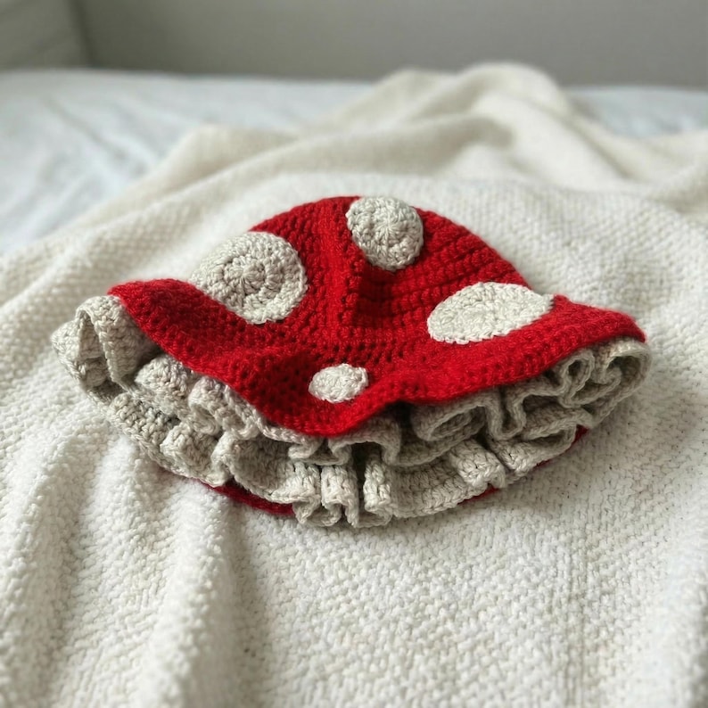 May include: A handmade crochet hat in the shape of a mushroom. The hat features a bright red cap with white spots and a ruffled, beige brim. The hat is designed to resemble a whimsical mushroom.