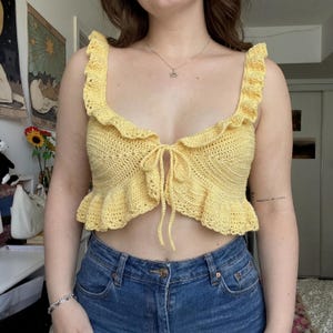 Op de afbeelding: Een gele gehaakte top met ruches en een striksluiting. De handgemaakte top heeft een open weefpatroon. Het kledingstuk is een crop top stijl, perfect voor warm weer.