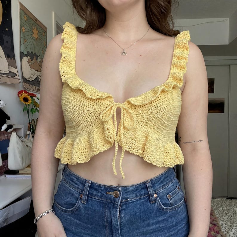 Op de afbeelding: Een gele gehaakte top met ruches en een striksluiting. De handgemaakte top heeft een open weefpatroon. Het kledingstuk is een crop top stijl, perfect voor warm weer.