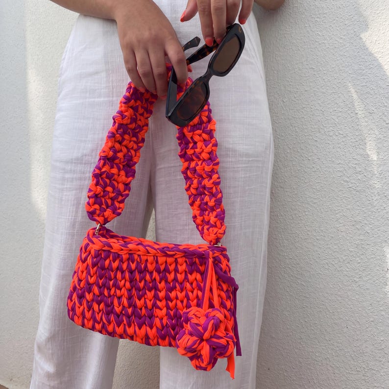 May include: A vibrant, handmade crochet bag in a striking orange and purple color scheme. The bag features a rectangular body with matching straps and a decorative pom-pom. Black sunglasses are held near the bag. The bag is a stylish accessory.
