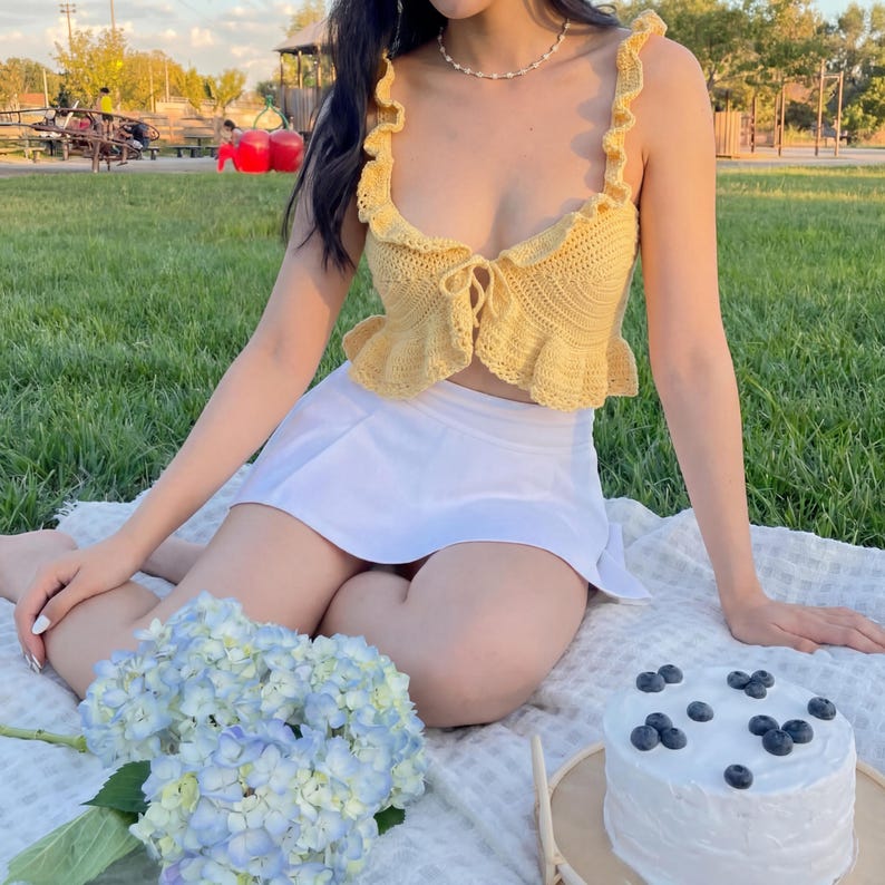 Op de afbeelding: Een gele gehaakte top met ruches en strikdetail, gecombineerd met een witte geplooide rok. De outfit wordt gedragen op een picknickkleed, met een boeket blauwe bloemen en een taart met blauwe bessen.