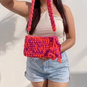 May include: A handmade, rectangular handbag in vibrant orange and purple. The bag features a braided strap and a decorative flower-shaped accent. The bag is made of a thick, woven material, and the colors are bold and eye-catching.