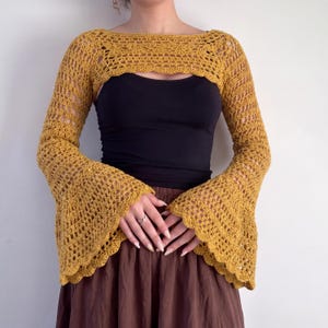 May include: A mustard yellow crochet shrug featuring long, flared bell sleeves with a scalloped edge and an intricate openwork pattern. The garment has a wide boat neckline and is worn over a plain black tank top and a solid brown skirt.