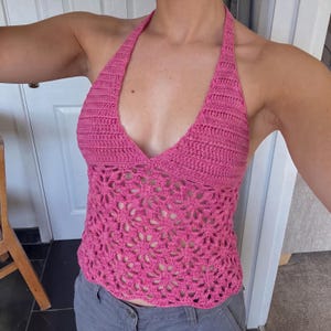 May include: A bright pink crochet halter top featuring a solid stitched upper bodice and a lower section crafted with an openwork floral pattern. The garment has a deep V-neckline and ties around the neck, showcasing a detailed yarn texture.