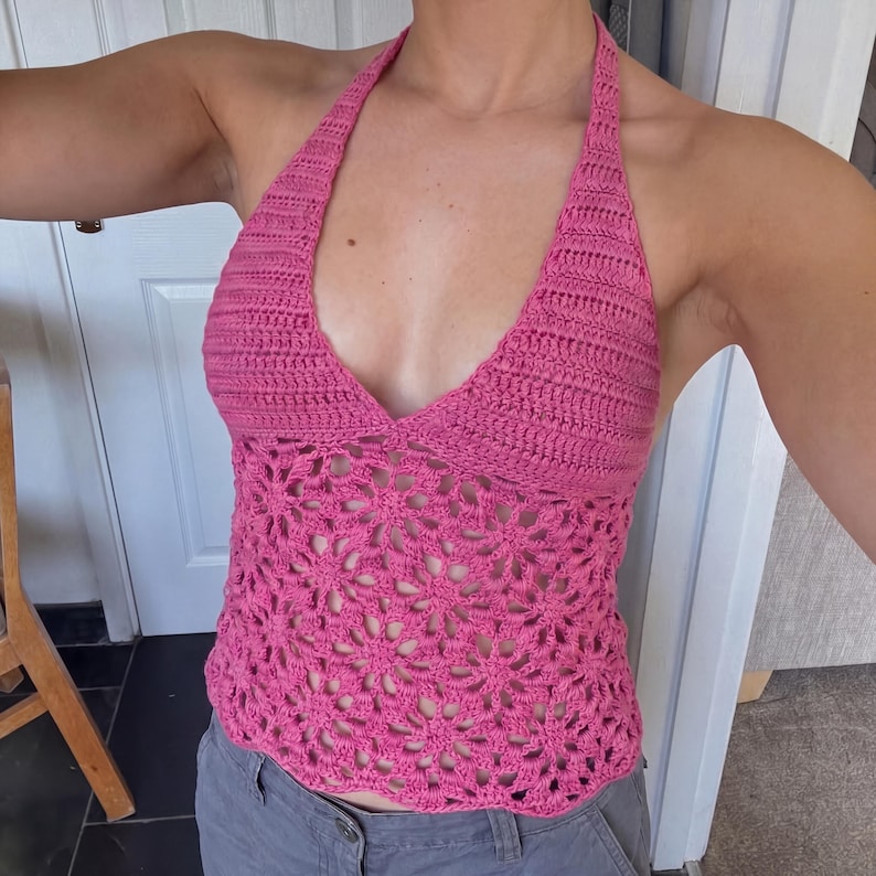 May include: A bright pink crochet halter top featuring a solid stitched upper bodice and a lower section crafted with an openwork floral pattern. The garment has a deep V-neckline and ties around the neck, showcasing a detailed yarn texture.