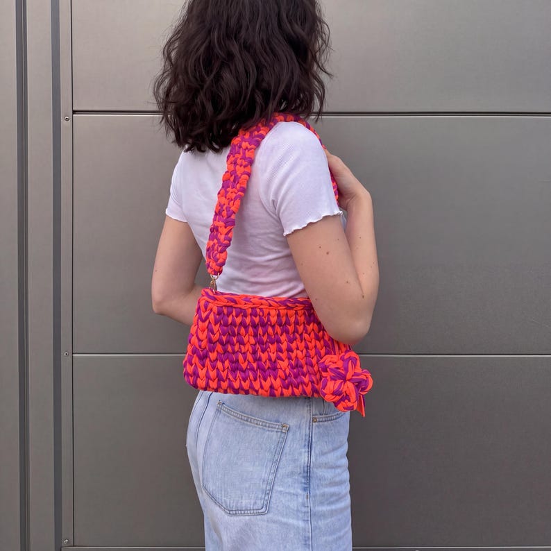 May include: A handmade, vibrant shoulder bag in a striking combination of neon orange and magenta. The bag features a thick, braided strap and a rectangular body, showcasing a bold, textured knit design. The bag is a stylish accessory.