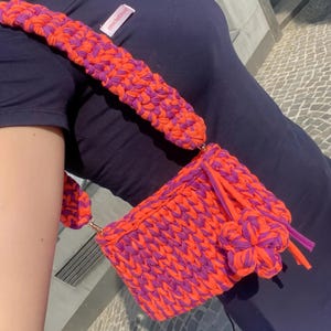 May include: A handmade, rectangular crochet bag in vibrant orange and purple. The bag features a matching strap and a decorative flower accent. The bag is made of thick, textured yarn and has a metal clasp. The text "crochetbyis" is visible on the strap.