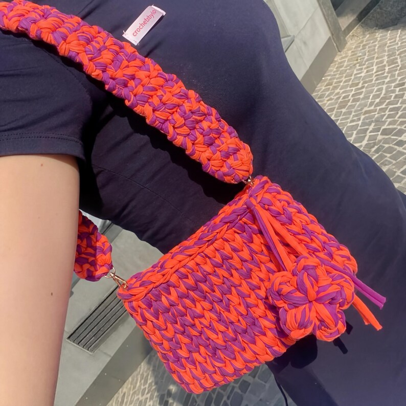 May include: A handmade, rectangular crochet bag in vibrant orange and purple. The bag features a matching strap and a decorative flower accent. The bag is made of thick, textured yarn and has a metal clasp. The text "crochetbyis" is visible on the strap.