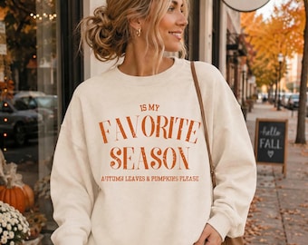 May include: A cream-coloured crewneck sweatshirt featuring orange distressed text that reads IS MY FAVOURITE SEASON and AUTUMN LEAVES & PUMPKINS PLEASE. The long-sleeved pullover has a ribbed neckline, cuffs, and hem, with a relaxed fit design.