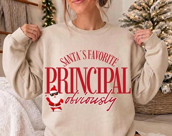 May include: A beige crewneck sweatshirt featuring red text that reads SANTA'S FAVORITE PRINCIPAL obviously. Below the text is a graphic of a cartoon Santa Claus wearing black sunglasses, a red hat, and a red suit with a black belt.