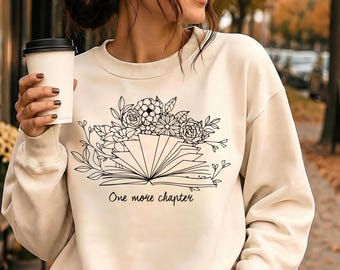May include: A cream-colored crewneck sweatshirt featuring a black line art graphic of an open book with a floral arrangement emerging from the pages. The text "One more chapter" is printed in a cursive script font below the book illustration.