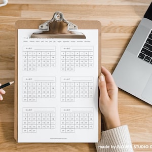 May include: A clipboard with a habit tracker calendar is held on a wooden surface. The calendar is printed in black ink and has boxes for tracking habits. A laptop and pencil are also visible.