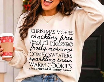 May include: A cream-colored crewneck sweatshirt featuring black text that reads: CHRISTMAS MOVIES, crackling fires, cold noses, frosty mornings, COMFY SWEATERS, sparkling snow, WARM COOKIES, gingerbread & candy canes in various fonts.