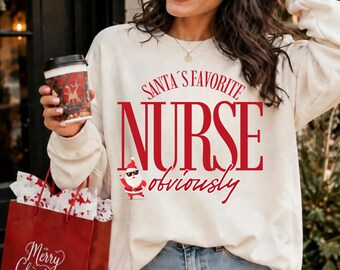 May include: A cream-colored crewneck sweatshirt featuring red text that reads "SANTA'S FAVORITE NURSE obviously" and a small graphic of Santa Claus wearing sunglasses. The sweatshirt has long sleeves and a ribbed neckline.