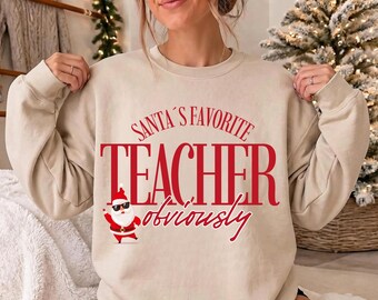 May include: A beige crewneck sweatshirt featuring red text that reads SANTA'S FAVORITE TEACHER obviously. Below the text is a graphic of Santa Claus wearing black sunglasses and a red suit, waving with one hand while standing in a pose.
