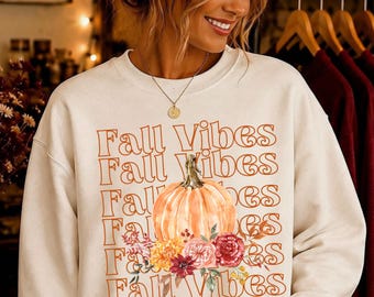 May include: A cream-colored crewneck sweatshirt featuring the repeating orange text Fall Vibes. The design includes a central illustration of a light orange pumpkin surrounded by a cluster of yellow, pink, and deep red flowers.
