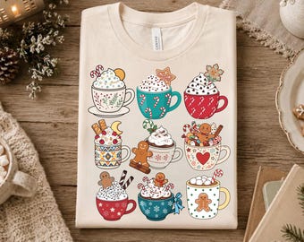 May include: A cream-colored t-shirt featuring a graphic print of nine different mugs filled with hot beverages, whipped cream, gingerbread cookies, candy canes, and festive holiday decorations in various colors including red, green, and blue.