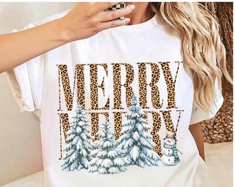 May include: A white short-sleeved t-shirt featuring the text MERRY MERRY in a brown and black leopard print pattern. Below the text are three snow-covered evergreen trees and a small snowman wearing a hat and a blue scarf.