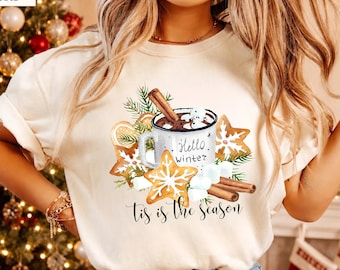 May include: A cream-colored t-shirt featuring a graphic of a mug labeled Hello Winter, surrounded by gingerbread cookies, marshmallows, and cinnamon sticks. The text 'tis is the season' is printed in black cursive script below the illustration.