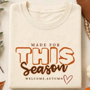May include: A cream-coloured t-shirt featuring the text "MADE FOR THIS Season" in brown and orange block and script lettering. Below the text, the words "WELCOME, AUTUMN" are printed in small brown letters next to a simple red heart outline.