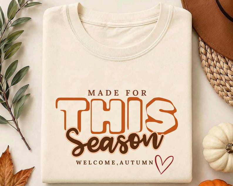 May include: A cream-coloured t-shirt featuring the text "MADE FOR THIS Season" in brown and orange block and script lettering. Below the text, the words "WELCOME, AUTUMN" are printed in small brown letters next to a simple red heart outline.