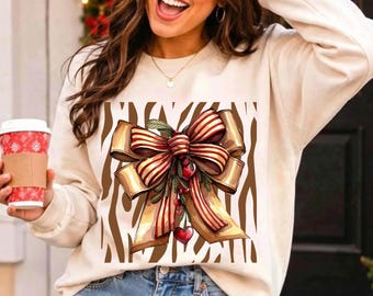 May include: A cream-colored crewneck sweatshirt featuring a central graphic print. The design displays a large, multi-layered bow with red and gold stripes, accented by small green leaves, red berries, and a heart, all set against a brown zebra print.