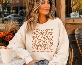 May include: A cream-coloured crewneck sweatshirt featuring a repeating pattern of orange hand-drawn heart outlines on the front. Below the heart design, the text reads Fall & Me, Happily Ever After in a cursive, orange-coloured script font.