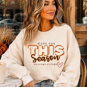 May include: A cream-coloured crewneck sweatshirt featuring the brown text "MADE FOR THIS Season" and "WELCOME, AUTUMN" with a small red heart outline. The garment has long sleeves and a ribbed neckline, cuffs, and hem, providing a casual fit.