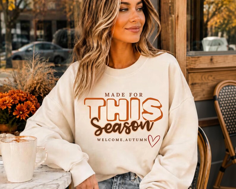 May include: A cream-coloured crewneck sweatshirt featuring the brown text "MADE FOR THIS Season" and "WELCOME, AUTUMN" with a small red heart outline. The garment has long sleeves and a ribbed neckline, cuffs, and hem, providing a casual fit.
