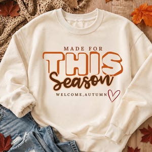 May include: A cream-coloured crewneck sweatshirt featuring the text MADE FOR THIS Season WELCOME, AUTUMN in orange and brown lettering, accompanied by a small red heart outline. The design is centred on the front of the long-sleeved garment.