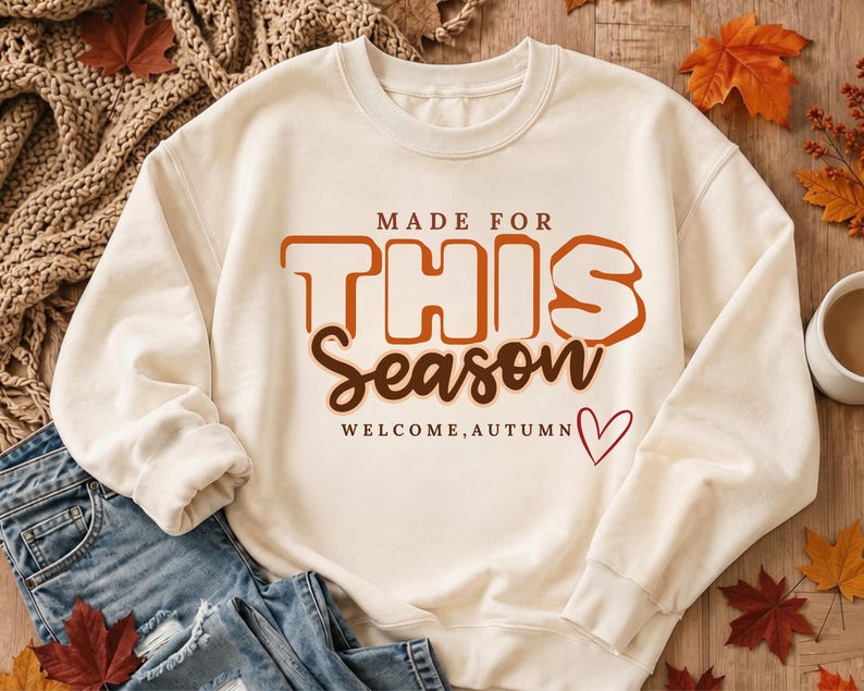 May include: A cream-coloured crewneck sweatshirt featuring the text MADE FOR THIS Season WELCOME, AUTUMN in orange and brown lettering, accompanied by a small red heart outline. The design is centred on the front of the long-sleeved garment.