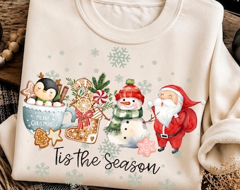 May include: A cream-colored crewneck sweatshirt featuring a festive graphic of a penguin in a mug, a gingerbread stocking, a snowman, and Santa Claus. The text "Merry Christmas" is on the mug, and "Tis the Season" is printed below.