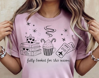 May include: A light pink t-shirt featuring a black graphic print of a stack of three books topped with a heart-patterned candle, a mug of hot cocoa with candy canes, and two airplane tickets, with the text fully booked for this season.