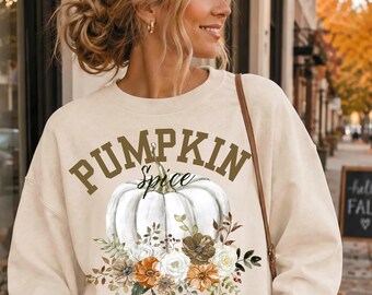 May include: A cream-colored crewneck sweatshirt featuring the text PUMPKIN SPICE in brown block letters. Below the text is a graphic of a white pumpkin surrounded by a floral arrangement of white, orange, and brown flowers and green leaves.