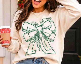 May include: A cream-colored crewneck sweatshirt featuring a large, dark green line-art graphic of a decorative bow with mistletoe sprigs and the text "Just Merry" written in a cursive script above the bow design.