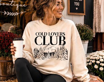 May include: A cream-coloured crewneck sweatshirt featuring the black text COLD LOVERS CLUB above a detailed line art illustration of a mountain range and a dense forest of pine trees. The long-sleeved garment has a ribbed collar and cuffs.