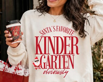 May include: A cream-colored crewneck sweatshirt featuring red text that reads SANTA'S FAVORITE KINDERGARTEN obviously. Below the word KINDER, there is a small graphic of Santa Claus wearing sunglasses and a red suit, positioned on the left side.