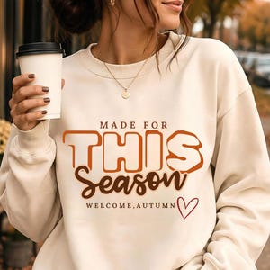May include: A cream-coloured crewneck sweatshirt featuring the text "MADE FOR THIS SEASON" in large orange block letters, followed by "Season" in brown script, and "WELCOME, AUTUMN" with a small red heart outline printed on the front.