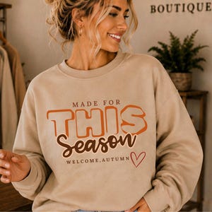 May include: A beige crewneck sweatshirt featuring the text MADE FOR THIS SEASON in orange and brown, with the additional text WELCOME, AUTUMN and a small red heart outline. The garment has long sleeves and a ribbed collar, cuffs, and hem.
