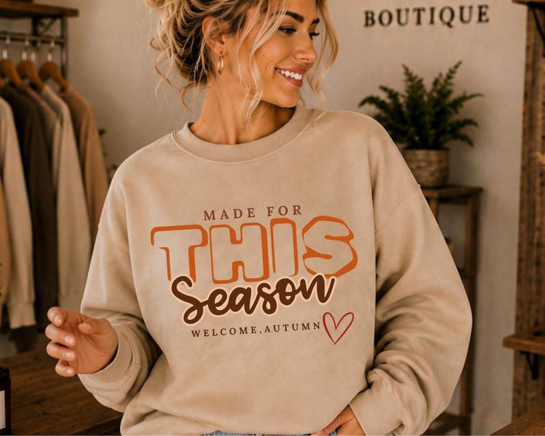 May include: A beige crewneck sweatshirt featuring the text MADE FOR THIS SEASON in orange and brown, with the additional text WELCOME, AUTUMN and a small red heart outline. The garment has long sleeves and a ribbed collar, cuffs, and hem.