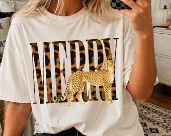 May include: An off-white short-sleeve t-shirt featuring the word MERRY in large, bold, black-outlined letters filled with a brown and black leopard print pattern. A realistic illustration of a standing cheetah is centered over the text.