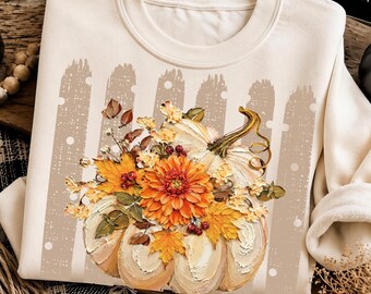 May include: A cream-colored crewneck sweatshirt featuring a graphic print of a white pumpkin adorned with a vibrant arrangement of orange dahlia flowers, maple leaves, and small red berries, set against a light brown picket fence design.