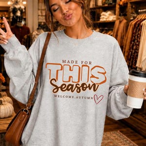 May include: A light grey crewneck sweatshirt featuring the orange and brown text MADE FOR THIS Season WELCOME, AUTUMN with a small red heart outline. The garment has long sleeves and a relaxed fit, worn with a brown shoulder bag and a white cup.
