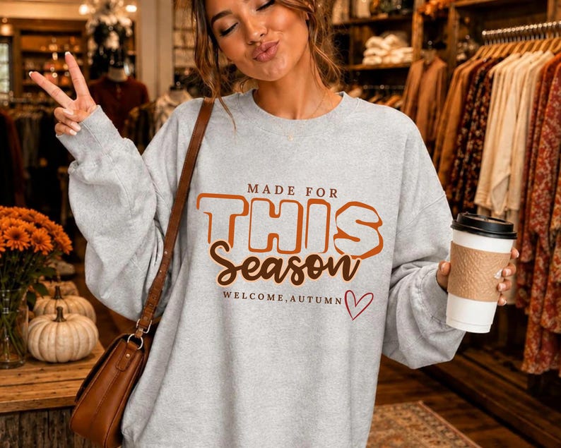 May include: A light grey crewneck sweatshirt featuring the orange and brown text MADE FOR THIS Season WELCOME, AUTUMN with a small red heart outline. The garment has long sleeves and a relaxed fit, worn with a brown shoulder bag and a white cup.