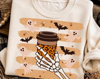 May include: A cream-colored sweatshirt featuring a graphic print of a skeleton hand holding a coffee cup with a leopard print sleeve. The design includes light orange horizontal brush strokes, small ghosts, black bats, and tiny star and dot accents.
