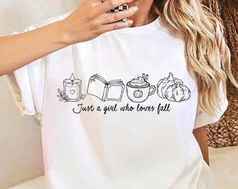 May include: A white t-shirt featuring a black line art graphic of a candle, an open book, a mug with whipped cream, and two pumpkins. Below the illustrations, the text reads, Just a girl who loves fall, in a cursive script font.