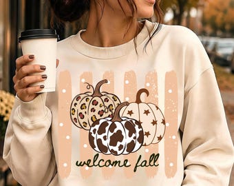 May include: A cream-colored crewneck sweatshirt featuring a graphic of three pumpkins with leopard, cow, and star patterns. The design includes vertical peach-colored brush strokes, white dots, and the text "welcome fall" in a dark brown script font.