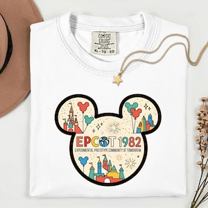 May include: White t-shirt with a colorful graphic of a Mickey Mouse head featuring a castle, balloons, and fireworks. The text "EPCOT 1982" and "EXPERIMENTAL PROTOTYPE COMMUNITY OF TOMORROW" are also displayed. A gold necklace with a star pendant is draped on the shirt.