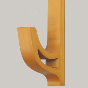 May include: A wall-mounted, mustard-yellow coat hook. The hook has a curved design with a cutout in the center, and a flat, rectangular back for mounting. The hook is made of a solid material, likely wood or a composite.