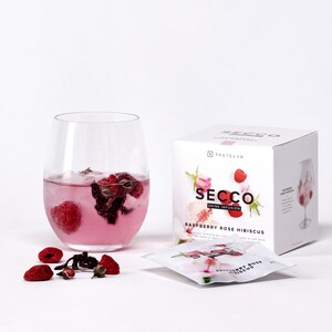 Könnte beinhalten: Ein Glas rosa Getränk mit Himbeeren und eine Schachtel mit "Secco Raspberry Rose Hibiscus" Getränkeinfusion. Das Getränk befindet sich in einem klaren, stielosen Glas. Die Schachtel und das Päckchen sind weiß mit rosa und roten Akzenten.